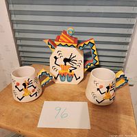 Full set of teapot and two mugs on wooden table showing whimsical dancer pattern and colorful geometric handles.