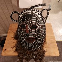 Front view of Zambian raffia cloth mask on wooden surface showing black, white, and brown linear patterns with fringed bottom and protrusions.