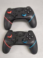 Front view of two black wireless game controllers with red and blue accents, showing buttons, joysticks, and directional pad.