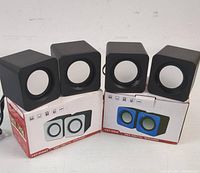 Four cube-shaped mini digital speakers arranged with two on top of two original retail boxes. Two speakers are black with blue fronts, two speakers are black with white fronts. Each speaker has a circular speaker cover visible.
