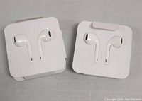 Two sets of generic white Apple-style wired earphones in original style plastic holders on a grey surface.