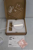 Open box showing the white circular LED base with attached USB cable, clear acrylic panel engraved with 1st anniversary heart and mouse design, instruction manual, and pink cloth inside foam packaging.