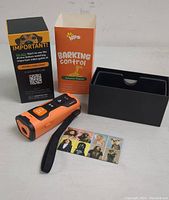 Photo of handheld orange and black WPS anti bark tool device, original packaging box, and instruction pamphlets laid out on a surface.
