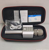 Closed-up photo of the Bluetooth microphone in the protective black and red carrying case alongside the USB charging cable, instruction manual, and quality certificate.