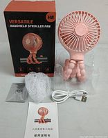 Handheld mini fan with flexible legs beside its box, USB cable, mesh cover, and manual on a flat surface.