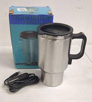 Photo of the stainless steel heated travel mug beside its original blue box. Mug has a black plastic handle and lid, and a detachable power cord with a 12V plug.