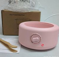 Pink circular hot towel steamer with timer dial, red indicator light, wooden tongs, and instruction manual displayed with the product box labeled 'Hot Towel Warmer'.