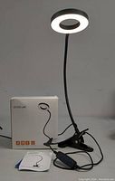 Photo of the assembled LED desk lamp powered on, showing its circular illuminated LED head, flexible neck, clamp base, and control cable.
