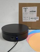 Photo of the round black mini electric turntable showing the on/off control switch and power connection port, placed next to the labeled box.