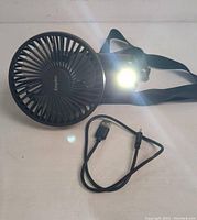 Front angle of black EasyAce mini fan with circular grill, USB charging cable beside it, and built-in light illuminated beside the fan grill, with adjustable strap attached.