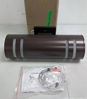 Front view of the cylindrical dark bronze outdoor light fixture with translucent horizontal bands, mounting bracket on back, mounting hardware, screws, anchors, and printed instructions on a white surface with cardboard box in background.