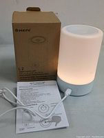 Photo showing Keni L2 smart touch lamp illuminated, connected with white USB cable, with instruction manual and box behind.