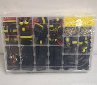 Top view of clear plastic organizer box with multiple compartments containing assorted tool kit accessories such as connector housings, sealing rings, metal pins, and wire clips in black, yellow, and red colors.