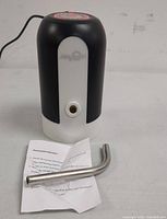 Front and side view of the black and white water dispenser pump with the metal curved spout and instruction sheet placed in front, showing water outlet nozzle and partial logo.