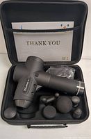 Open carrying case showing Wattne W1 massage gun with six different massage heads, USB charging cable, and a thank you card inside mesh pocket.