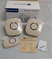 Photo showing two wireless receivers, one transmitter button, mounting screws in plastic bag, instruction manual, and product box with SadoTech branding and wireless doorbell labeling.