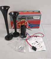 Full view of Horn Tech truck horn set including two long black trumpet-style horns, wiring, compressor unit, mounting hardware in plastic bag, instruction page, and red-gray retail box.