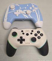 Front view of two wireless game controllers: a light blue and white controller with white buttons, and a white and black controller with colored rings on analog sticks.