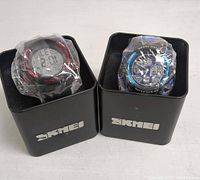 Front view of two watches in black SKMEI metal tins, one digital with red accents, one analog digital with blue accents
