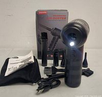 Front view of electric air duster with LED light on, showing main unit, multiple nozzle attachments, USB cable, user manual, and carrying pouch.