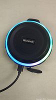Front view of Kunodi Bluetooth shower speaker showing illuminated LED ring around speaker grill and USB charging cable connected.