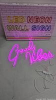 Pink illuminated LED neon wall sign spelling 'Good Vibes' laid flat with power cord and switch, packaging box in background with colorful 'LED NEON WALL SIGN' text.