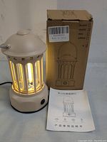 Portable beige camping lantern glowing warm yellow light, next to original cardboard box and instruction manual with foreign text.