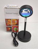 Image of the sunset lamp next to its box, showing the black metallic design with a round base and lens head. USB cable attached and packaging indicating 'Sunset lamp'.