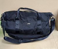 Front view of the black travel duffel bag with brand logo, handles, and shoulder strap visible.