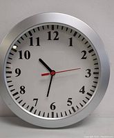 Front view of a round wall clock with silver frame, white face, black numerals, black hour and minute hands, and red second hand.