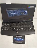 Photo showing black KW213 wired keyboard with USB plug, displayed in front of original packaging that highlights colored backlight feature