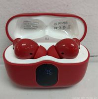 Red wireless earbuds inside an open charging case showing LED battery display and regulatory information on the inside lid.