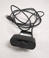 Front view of a black rectangular USB webcam with the lens covered and attached to a long black USB cable lying on a white surface.