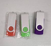 Three USB flash drives in orange, green, and purple with metal swivel covers, two sealed in plastic wrap, one unwrapped.