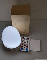 White egg-shaped baby night light on beige base next to cardboard box, face stickers, and user manual.