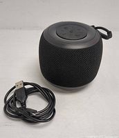 Black Bluetooth portable speaker with rounded mesh body and circular top control panel. USB charging cable beside it on a white surface.