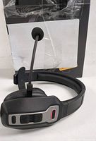 Left side close-up of the black Bluetooth wireless headset showing microphone boom and control buttons on ear cup, with original box behind.