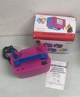 Photo of the pink electric balloon pump with blue handle placed on a surface with power cord and instruction manual, alongside the product box.