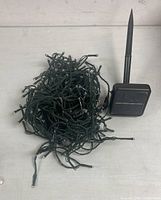 Photo of coiled green wire string Christmas lights beside a small black solar panel with stake for ground installation.