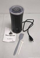 Electric SS-001 milk frother with power cord, user manual, gray silicone spatula, and white cleaning brush on flat surface.