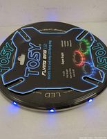 Photo of the black circular Tosy Flying Ring with blue LED lights illuminated on the ring's perimeter, showing the light effect and the shape.