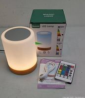 Photo showing Magic Light LED lamp illuminated, with its box, remote control and USB charging cable placed nearby on a flat surface.