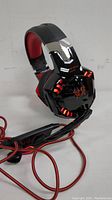 Side-view image showing over-ear headphones with black and red design, illuminated accents, and spider logo on ear cup. Wired connection with microphone and volume control visible.