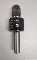 Wireless karaoke microphone front view showing metallic mesh head, control panel with buttons, and metallic handle against a plain background.