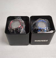 Two kids digital watches in black metal SKMEI boxes, one with red accents and one with blue accents, shown from the top with plastic protective wrap