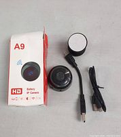 Photo of A9 Battery IP Camera, USB cable, short adapter cable, and round charging adapter with open box packaging