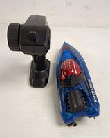 Remote control and Water Spider toy boat shown from angled top and side views. Boat is blue with red cockpit and spider web graphics. Remote is black with large dial.