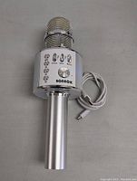 Front view of silver Bluetooth karaoke microphone showing control panel with labeled buttons and rotary dials. USB charging cable coiled beside microphone.