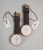 Two Brigada wristwatches side by side on a flat surface, showing face and leather strap with tags attached.