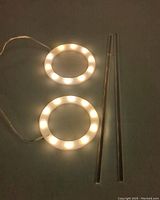 Two illuminated circular white LED rings side by side on dark surface with two clear acrylic stands laid adjacent.